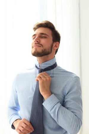 Photo for Morning dress up. Handsome young man in blue shirt adjusting his necktie and looking away. - Royalty Free Image