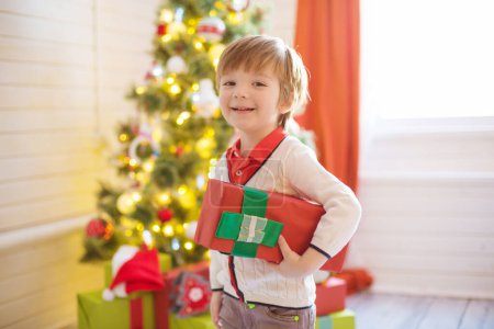Téléchargez les photos : Petit garçon tenir une boîte cadeau près d'un arbre de Noël décoré à la maison - en image libre de droit