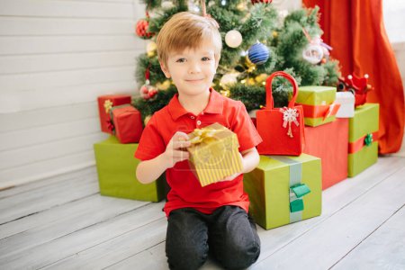 Téléchargez les photos : Mignon petit garçon d'environ cinq ans avec un cadeau dans une chambre de Noël décorée avec un arbre de Noël. - en image libre de droit