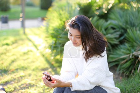 Foto de Chica coreana sentada en el parque y usando smartphone. - Imagen libre de derechos