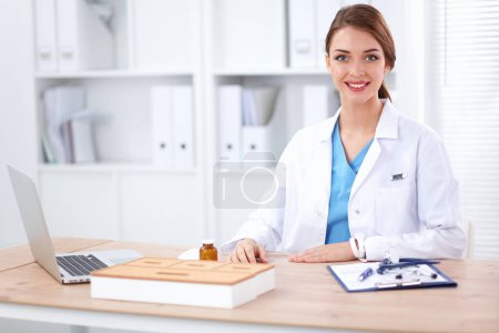 Foto de Hermosa joven sonriente doctora sentada en el escritorio y escribiendo. - Imagen libre de derechos