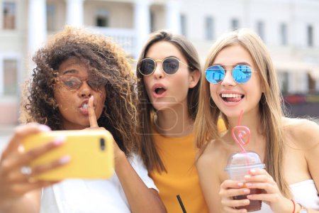 Photo for Three trendy cool hipster girls, friends drink cocktail and taking selfie in city background. - Royalty Free Image