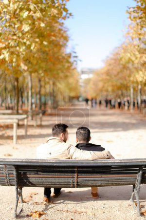 Téléchargez les photos : Vue arrière de deux gays câlins assis sur un banc dans un parc d'automne. - en image libre de droit