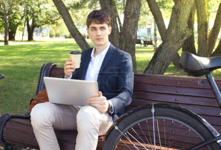 Téléchargez les photos : Un homme d'affaires en pause café. Il est assis sur un banc et travaille sur un ordinateur portable, à côté du vélo
. - en image libre de droit