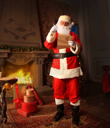 Téléchargez les photos : Père Noël debout dans sa chambre à la maison près de l'arbre de Noël et grand sac et lecture lettre de Noël ou liste de souhaits
. - en image libre de droit