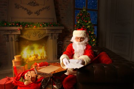 Foto de Santa sentado en el árbol de Navidad, sosteniendo cartas de Navidad y descansando junto a la chimenea
. - Imagen libre de derechos