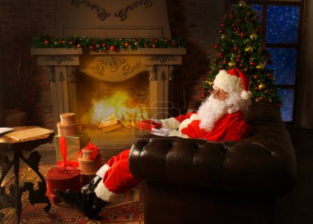 Foto de Retrato de Papá Noel feliz sentado en su habitación en casa cerca del árbol de Navidad y leyendo la carta de Navidad o la lista de deseos. - Imagen libre de derechos