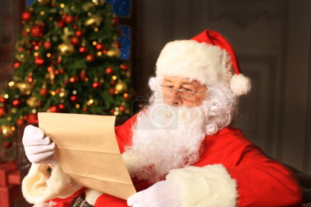 Téléchargez les photos : Portrait du Père Noël heureux assis dans sa chambre à la maison près de l'arbre de Noël et lisant la lettre de Noël ou la liste de souhaits. - en image libre de droit