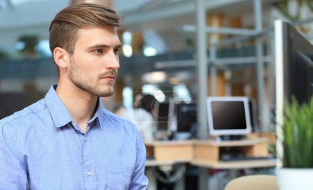 Téléchargez les photos : "Jeune homme assis et regardant l'écran d'ordinateur tout en travaillant au bureau
." - en image libre de droit