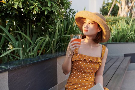 Téléchargez les photos : Belle asiatique fille assis dans le banc et boire du jus sous le soleil d'été - en image libre de droit