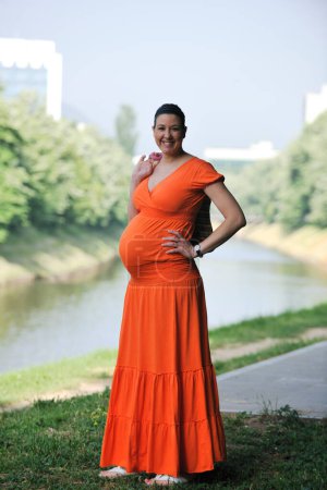 Photo for Pregnant woman in an orange dress posing outdoors, happy pregnancy - Royalty Free Image