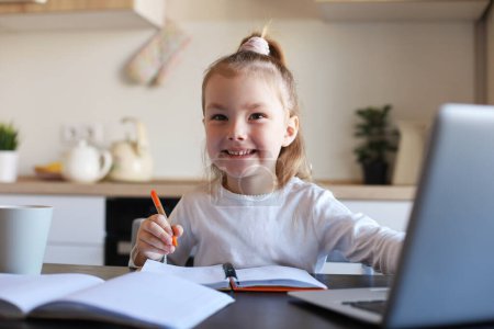 Téléchargez les photos : Souriant petite fille écriture étude en ligne à l'aide d'un ordinateur portable à la maison, mignon heureux petit enfant prendre Internet leçon web ou classe sur PC. - en image libre de droit