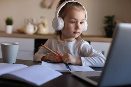 Foto de Sonriente niña en los auriculares estudio de escritura a mano en línea utilizando el ordenador portátil en casa, lindo niño pequeño feliz en los auriculares tomar lección web de Internet o clase en PC. - Imagen libre de derechos