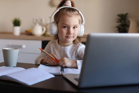Foto de Sonriente niña en los auriculares estudio de escritura a mano en línea utilizando el ordenador portátil en casa, lindo niño pequeño feliz en los auriculares tomar lección web de Internet o clase en PC. - Imagen libre de derechos