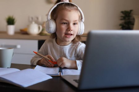 Téléchargez les photos : Souriant petite fille dans les écouteurs écriture étude en ligne à l'aide d'un ordinateur portable à la maison, mignon petit enfant heureux dans les écouteurs prendre des leçons Web Internet ou cours sur PC. - en image libre de droit