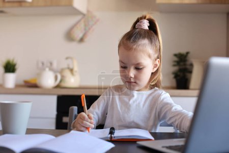 Téléchargez les photos : Sérieux petite fille écriture étude en ligne à l'aide d'un ordinateur portable à la maison, mignon heureux petit enfant prendre Internet leçon web ou classe sur PC. - en image libre de droit