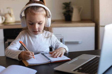Photo for Little girl in headphones sit at desk writing in notebook studying online do exercises at home, little child handwrite prepare homework on quarantine, have web class or lesson indoors. - Royalty Free Image