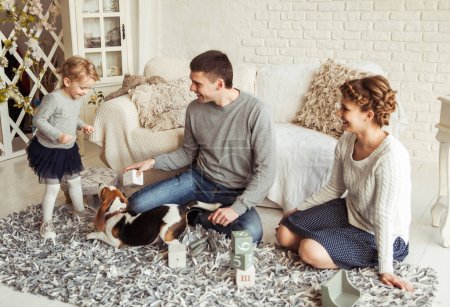 Téléchargez les photos : Famille heureuse jouer avec un chien de compagnie dans le salon spacieux - en image libre de droit