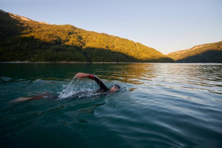 Téléchargez les photos : Athlète de triathlon nageant sur le lac au lever du soleil portant une combinaison - en image libre de droit