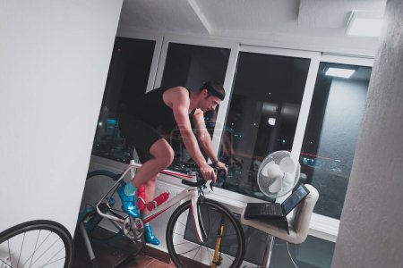 Foto de Hombre ciclismo en el entrenador de la máquina que está ejercitando en el hogar por la noche jugando juego de carreras de bicicletas en línea - Imagen libre de derechos