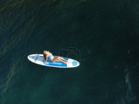 Photo for "Aerial drone view on young attractive brunette woman with long hair in blue swimsuit, swimming on sup around volcanic rocks, like in Iceland. Summer holiday vacation and travel concept." - Royalty Free Image