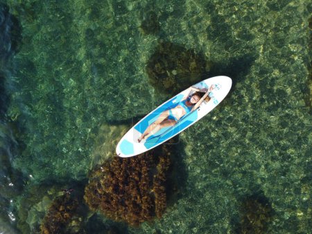 Foto de "Vista aérea del dron sobre una joven atractiva morena con el pelo largo en traje de baño azul, nadando en sup alrededor de rocas volcánicas, como en Islandia. Vacaciones de verano y concepto de viaje." - Imagen libre de derechos