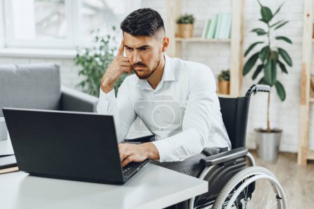 Téléchargez les photos : "Homme concentré sérieux en fauteuil roulant utilisant son ordinateur portable pour le travail / à la recherche d'un emploi dans Internet" - en image libre de droit