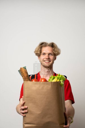 Téléchargez les photos : Portrait de livreur heureux en uniforme rouge souriant tout en portant un sac en papier avec des produits alimentaires isolés sur fond blanc. - en image libre de droit