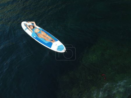 Foto de "Vista aérea del dron sobre una joven atractiva morena con el pelo largo en traje de baño azul, nadando en sup alrededor de rocas volcánicas, como en Islandia. Vacaciones de verano y concepto de viaje." - Imagen libre de derechos