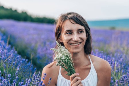 Photo for "Close up portrait of happy young brunette woman in white dress on blooming fragrant lavender fields with endless rows. Warm sunset light. Bushes of lavender purple aromatic flowers on lavender fields." - Royalty Free Image