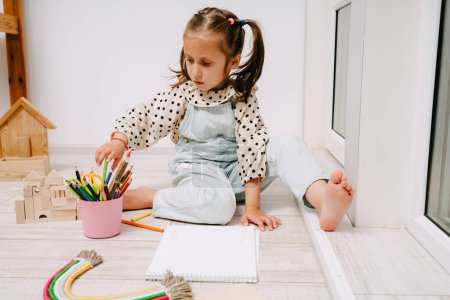 Foto de Niña se sienta en el suelo y dibuja en un cuaderno de bocetos con lápices de colores. - Imagen libre de derechos