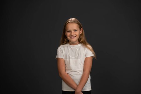 Photo for Smiling girl looking timid or frustrated at the camera wearing white t-shirt isolated on dark grey or black background. Child emotions concept - Royalty Free Image