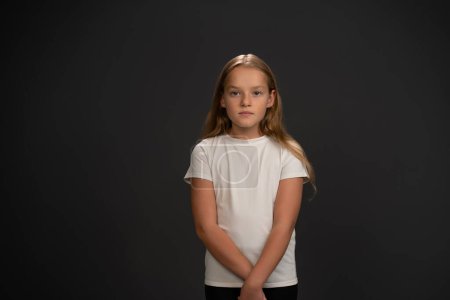 Foto de Niña de pie mirando con interrogación o frustrada a la cámara con una camiseta blanca aislada sobre fondo gris oscuro o negro - Imagen libre de derechos