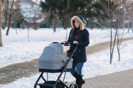 Photo for Beautiful mother walking in the park with her little baby in stoller. Woman dressed in blue jaket with hood and jeans. Warm boots - Royalty Free Image