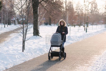 Foto de "Hermosa madre caminando en el parque con su pequeño bebé en Stoller. Mujer vestida con jaket azul con capucha y jeans. Botas calientes
" - Imagen libre de derechos