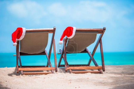 Téléchargez les photos : "Chapeau de Père Noël sur chaise longue avec eau de mer turquoise et sable blanc. Concept vacances de Noël
" - en image libre de droit