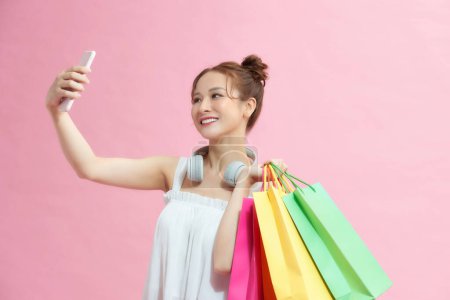 Téléchargez les photos : Fille asiatique excitée shopaholic utilisant téléphone posant avec des sacs à provisions sur fond rose. Vente et remises saison - en image libre de droit