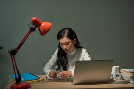 Foto de Estudiante bastante joven sentada en el escritorio y haciendo su tarea, se está conectando a Internet con un ordenador portátil - Imagen libre de derechos