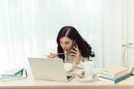 Photo for "Confident smiling attractive young asian woman entrepreneur having lunch break while sitting at the office desk and using mobile phone" - Royalty Free Image