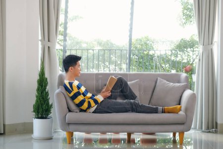 Téléchargez les photos : Young man reading book while sitting on the couch - en image libre de droit