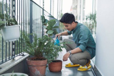 Téléchargez les photos : Attrayant jeune homme sur appartement balcon plantes d'arrosage en boîte de Blue Watering Can - en image libre de droit