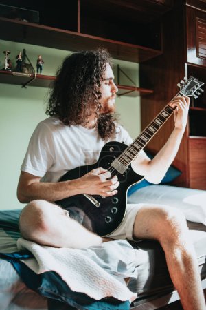 Téléchargez les photos : "Jeune homme aux cheveux longs jouant de la guitare électrique dans sa chambre. Chemise blanche assise sur le lit confortable dans la chambre et pensant à quelque chose. Éducation musicale concept image." - en image libre de droit