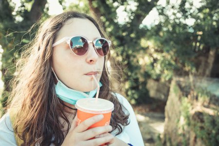 Téléchargez les photos : "Une jeune femme boit un milk-shake avec des lunettes de soleil et un masque pendant une journée ensoleillée, une plage ensoleillée et un concept de vacances" - en image libre de droit