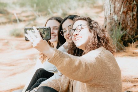 Téléchargez les photos : "Three young woman student taking a selfie in the forest during a sunny day, friendship concept, love" - en image libre de droit