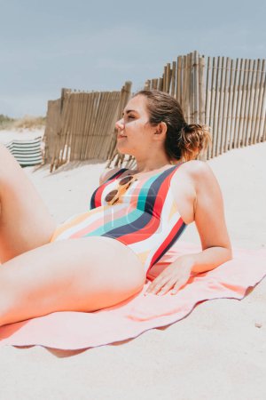 Foto de "Young woman looks away from camera while taking a sun bath wearing a colorful swimsuit at the beach, travel young holiday concept, copy space, social network, sunglasses user concept" - Imagen libre de derechos