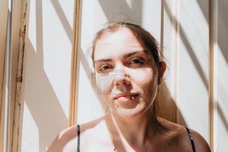 Téléchargez les photos : "Close up of a young woman looking to camera next to a window during a super sunny day, copy space, reflexion and thinking concept, sad and anxiety, self care" - en image libre de droit