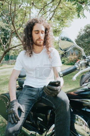 Foto de "Young long hair motorbike guy checking his phone while sitting on his old school motorbike during a break from the road route. Liberty life, young man heavy metal, white tshirt and gloves." - Imagen libre de derechos