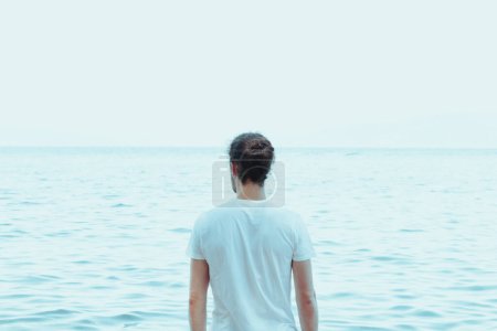 Téléchargez les photos : Young hipster male with long hair and a bun looking to the ocean, during a sunny day, reflection and yoga concept, serenade, copy space - en image libre de droit