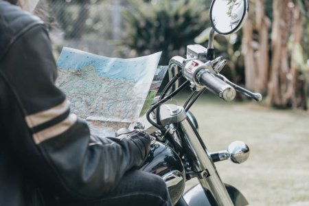 Foto de Close up of a man in a motorbike checking a map while doing a road trip. Travel concept. Gloves and rude hands holding a map over a old school motorbike. Copy space. - Imagen libre de derechos
