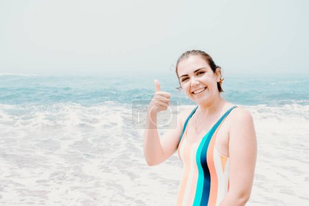 Foto de "Mujer joven en traje de baño saluda a la cámara durante un día soleado en la playa con el océano como fondo, libertad y concepto de vacaciones, espacio para copiar" - Imagen libre de derechos